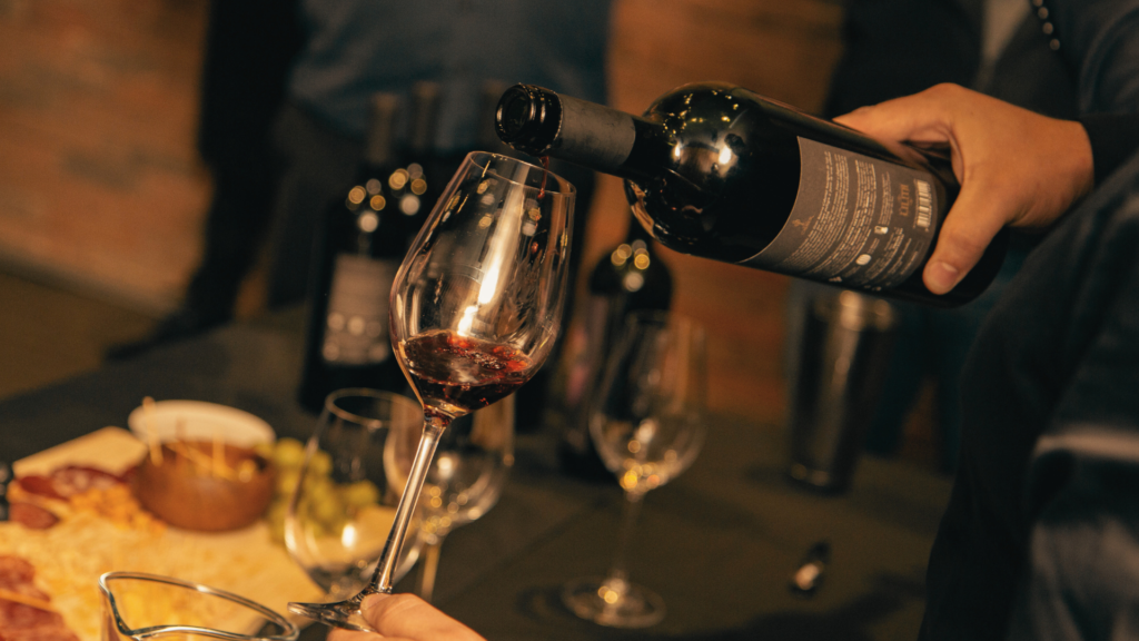 Mão segurando uma garrafa de vinho enquanto serve uma taça em um evento de degustação, com uma mesa ao fundo repleta de queijos e embutidos para harmonização.