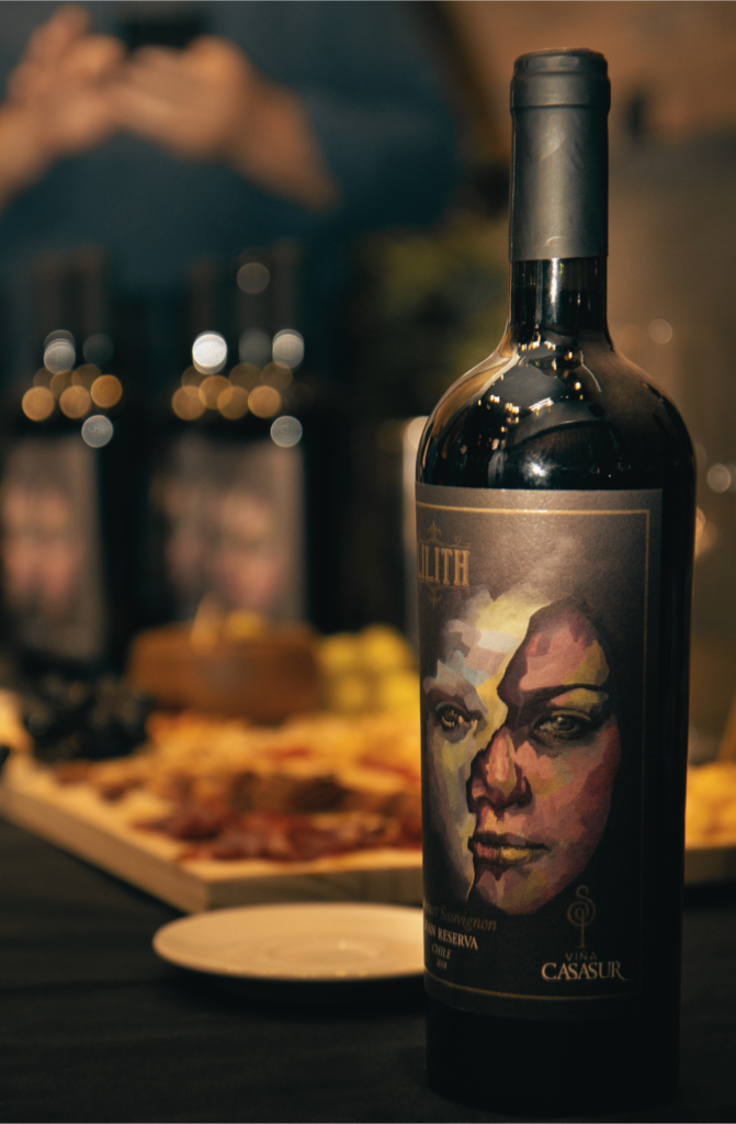 Garrafa de vinho Lilith Grand Reserva em foco, com um rótulo artístico vibrante, exibida em uma mesa de degustação com queijos e frios ao fundo.